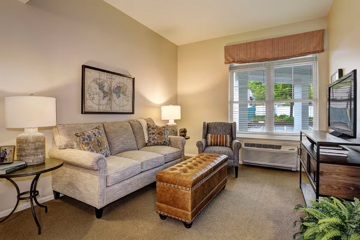 A cozy living room with a beige sofa adorned with patterned cushions, a brown leather tufted ottoman in front, a gray armchair with a striped pillow, two table lamps on either side of the sofa, a framed world map on the wall, a large window with blinds and a valance, a flat-screen TV on a wooden stand, and a green potted plant in the corner.