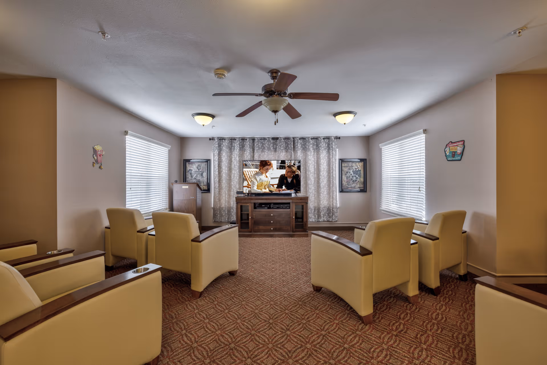 A cozy living room with six beige armchairs arranged in two rows facing a flat-screen TV on a wooden stand. The room has two windows with blinds, framed pictures on the walls, a ceiling fan, and patterned carpet flooring.