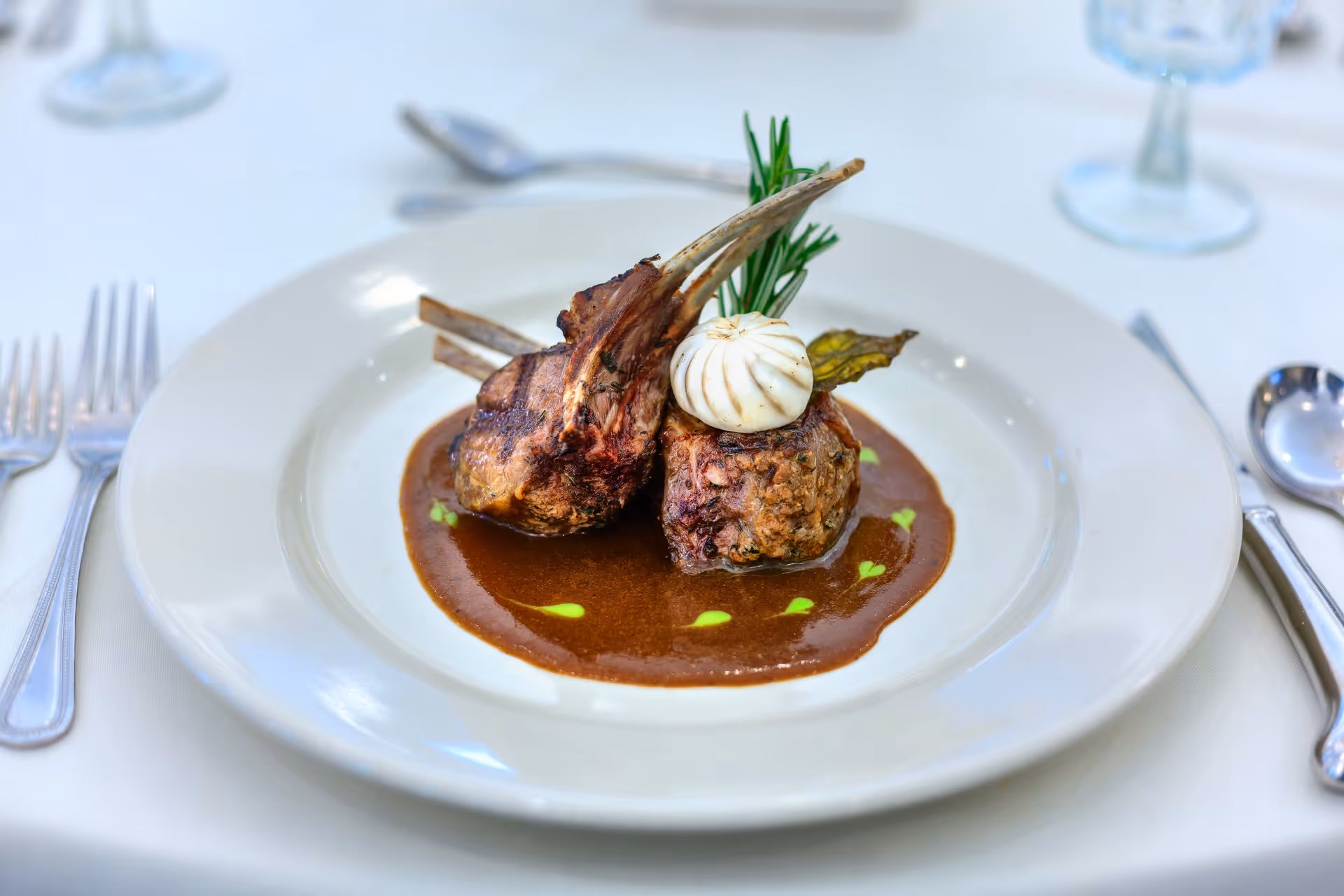A plated gourmet lamb chop entrée with sauce and garnishes on a white table setting.