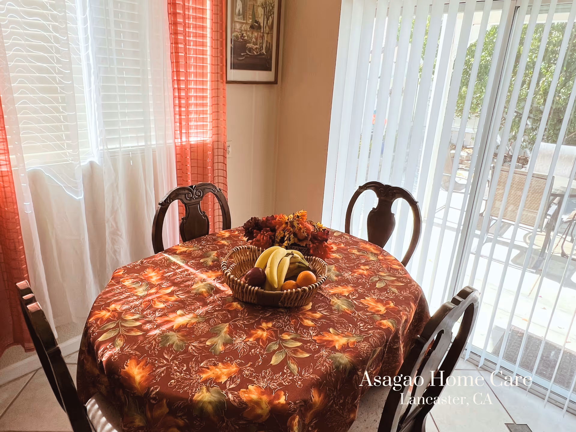 A dining area with a round table covered by a floral autumn-themed tablecloth. On the table is a basket filled with bananas, oranges, and other fruits, along with a decorative flower arrangement. Four wooden chairs surround the table. The room has large windows with white and red curtains on one side and vertical blinds on the other, allowing natural light to enter. Outside the window, patio furniture and greenery are visible.
