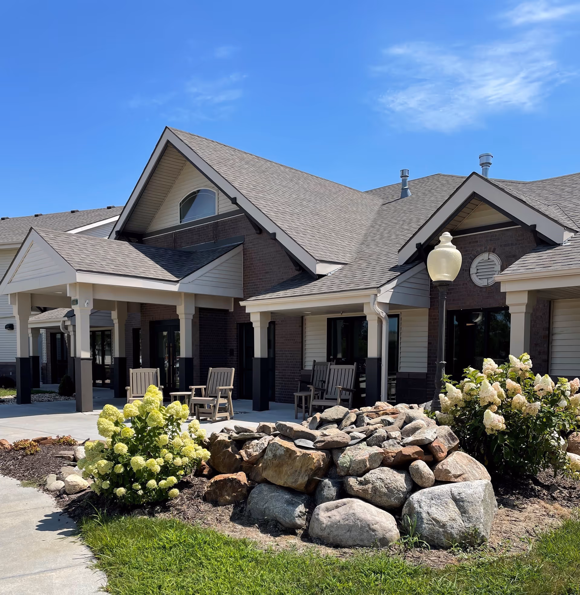 Exterior view of Valley Lakes Assisted Living facility showing a building with a peaked roof, a covered entrance, outdoor seating with wooden chairs, landscaped garden with flowering bushes and a rock arrangement, under a clear blue sky.