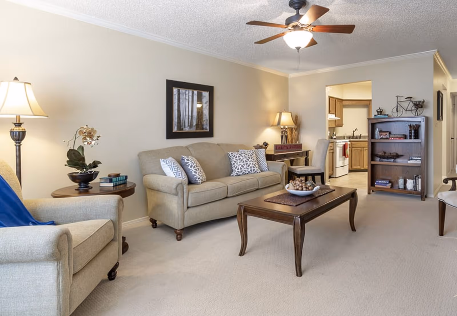 A cozy living room with beige walls and carpet, featuring a beige sofa with patterned cushions, a matching armchair with a blue throw, a wooden coffee table with a decorative bowl, a side table with a lamp and plant, a wooden bookshelf with books and decor, and a ceiling fan with a light. The room opens into a kitchen area with wooden cabinets and white appliances.