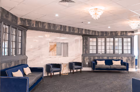A spacious lounge area with dark blue sofas and armchairs arranged around a carpeted floor. The walls have a combination of marble and dark wood paneling with large windows. The ceiling features recessed lighting and decorative chandeliers.