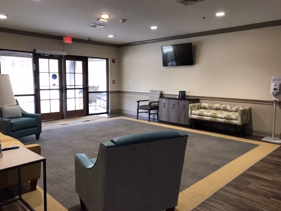 A spacious common area with a large carpet, several chairs including a teal armchair and a patterned loveseat, a wooden cabinet with a flat-screen TV mounted above it, and glass double doors leading outside. The room has neutral-colored walls and ceiling lights, with a hand sanitizer dispenser near the wall.