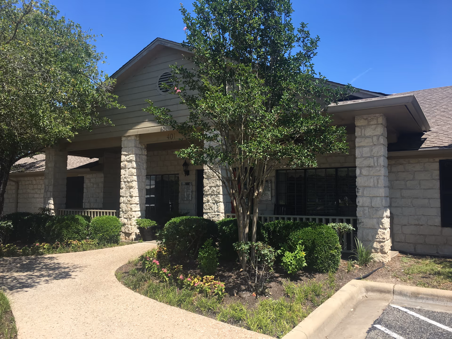 Stone-faced single-story building entrance with columns, a curved walkway, and landscaping under a clear blue sky.
