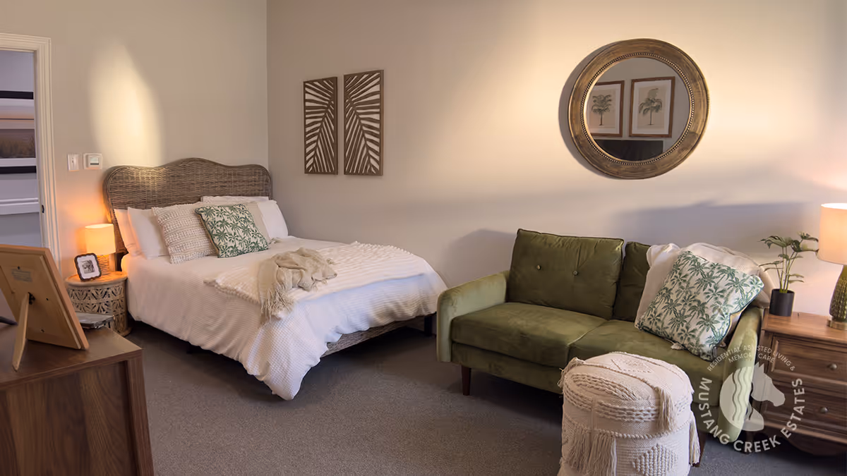 A cozy bedroom featuring a bed with white bedding and decorative pillows, a green loveseat with patterned pillows, a round mirror on the wall, two framed botanical prints, a wooden nightstand with a lamp and plant, and a small round ottoman in front of the loveseat.