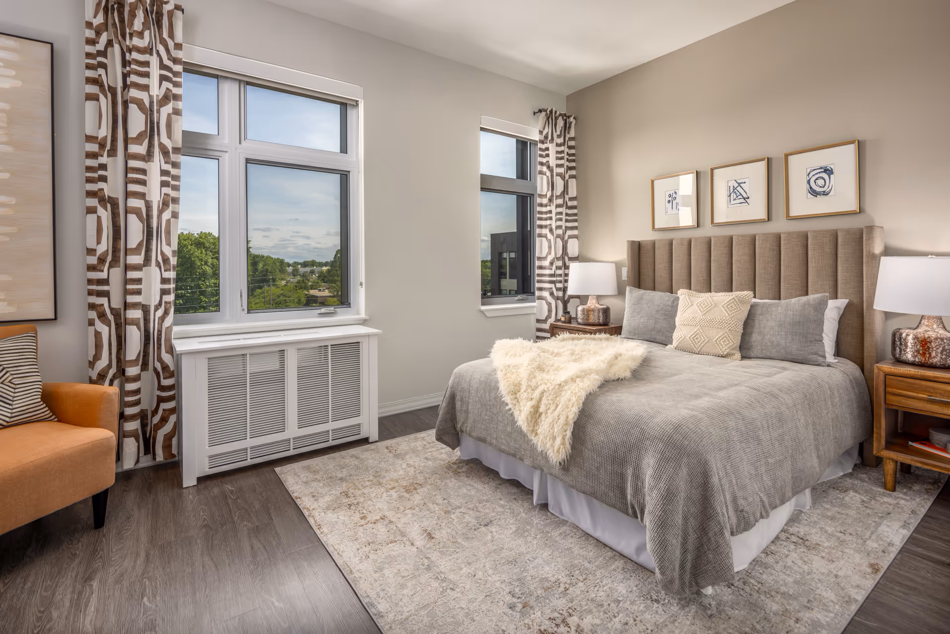A well-lit bedroom with a large bed featuring a beige upholstered headboard, gray bedding, and decorative pillows. There are two wooden nightstands with lamps on either side of the bed. The room has two windows with patterned curtains, a light-colored area rug, and a comfortable orange armchair in the corner. The walls are painted light beige and decorated with three framed abstract artworks above the bed.