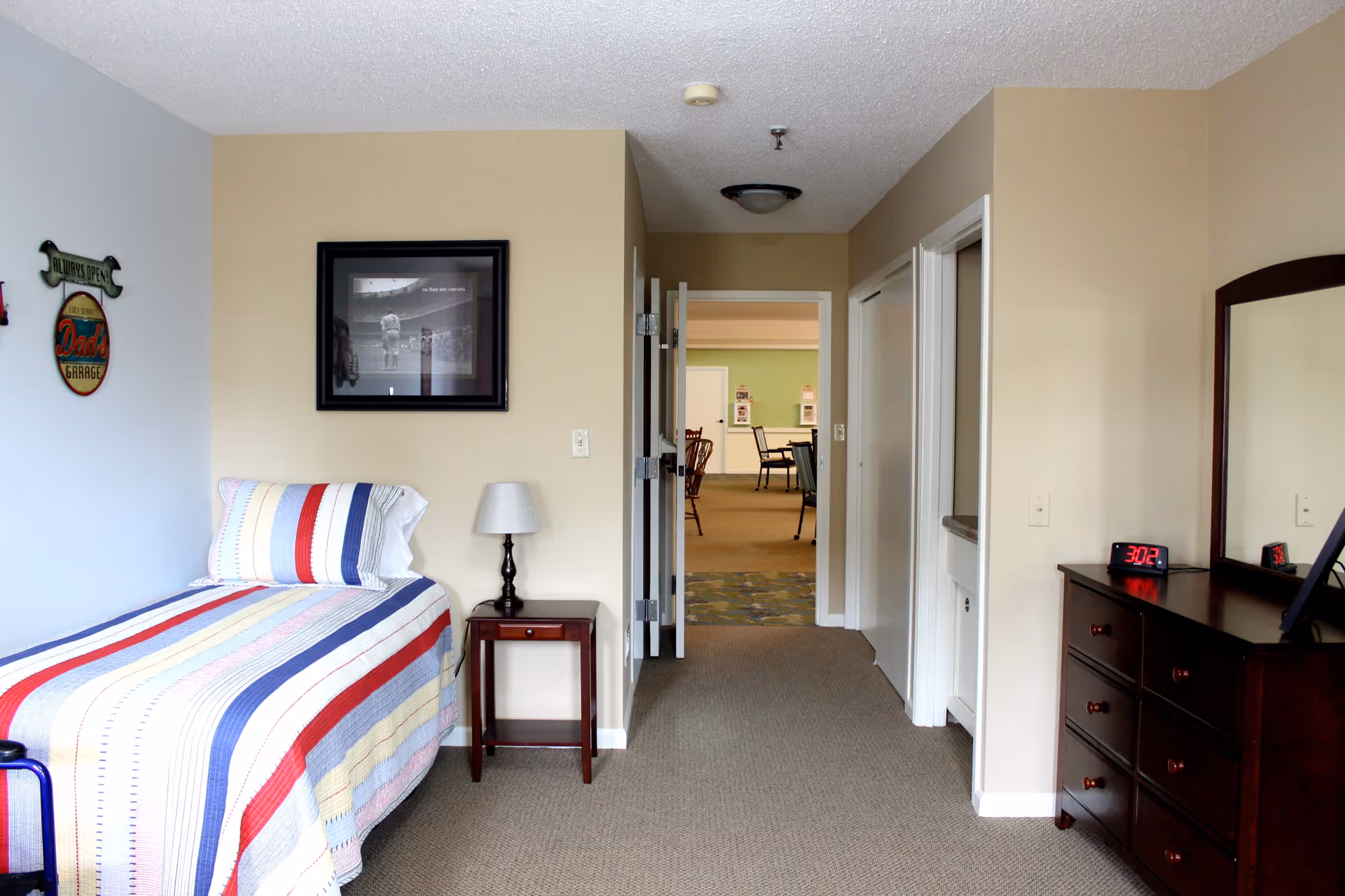 A senior living facility bedroom with a single bed covered in a striped quilt with red, blue, yellow, and white colors. Next to the bed is a small wooden nightstand with a lamp. On the wall above the bed are decorative signs and a framed black and white photo. Across from the bed is a dark wooden dresser with a mirror and a digital clock showing 3:02. The room has beige walls and carpeted floor. An open door leads to a common area with tables and chairs visible in the background.
