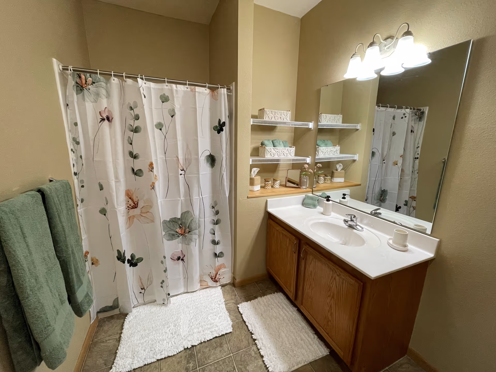 A bathroom with a floral shower curtain, two green towels hanging on a wall rack, a white sink countertop with a wooden cabinet underneath, a large mirror above the sink, and shelves holding neatly arranged towels and decorative items.