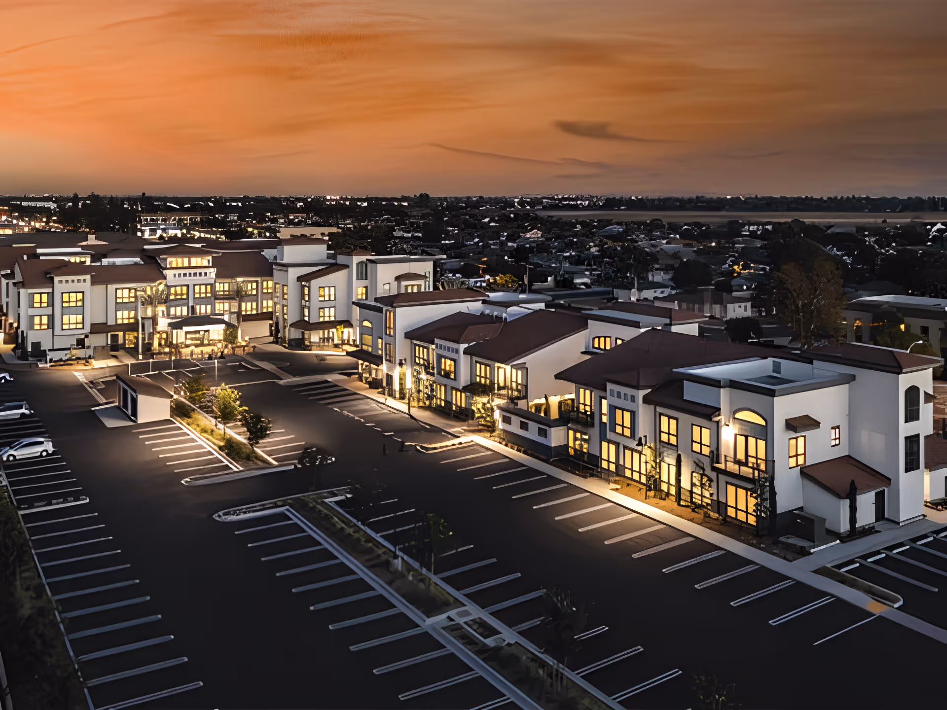Aerial view of Westmont of Cypress senior living facility at dusk, showing a large, modern building complex with illuminated windows and empty parking lots under a colorful sunset sky.