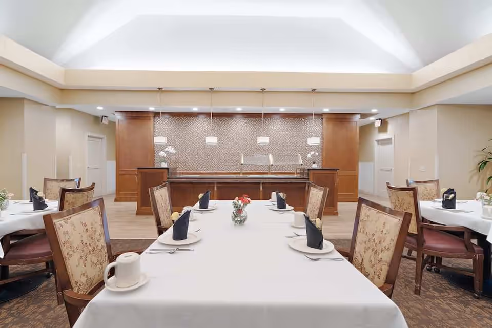 Bright dining room with tables set with white tablecloths, folded napkins, chairs, and a serving counter at the far wall.