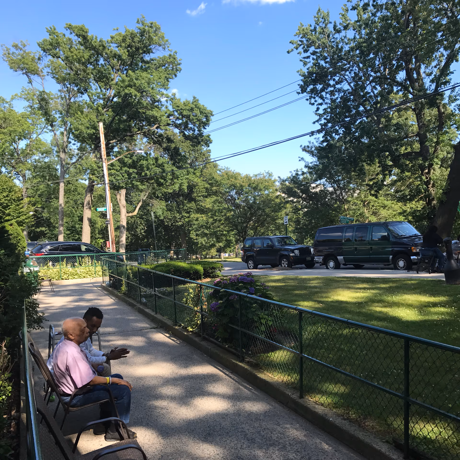 Two men sitting on chairs along a paved walkway next to a green chain-link fence with trees and parked vehicles in the background under a clear blue sky.
