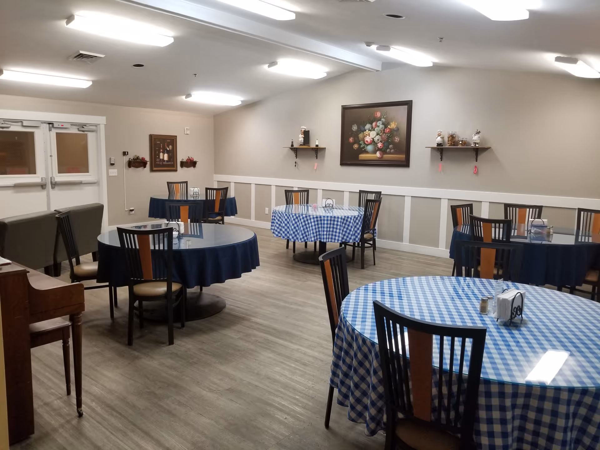 Interior view of a dining room with several round tables covered with blue and blue-checkered tablecloths, each surrounded by chairs. The room has light-colored walls with decorative molding, a painting of flowers on the wall, shelves with small decorative items, and overhead fluorescent lighting. There is a piano on the left side and double doors in the background.