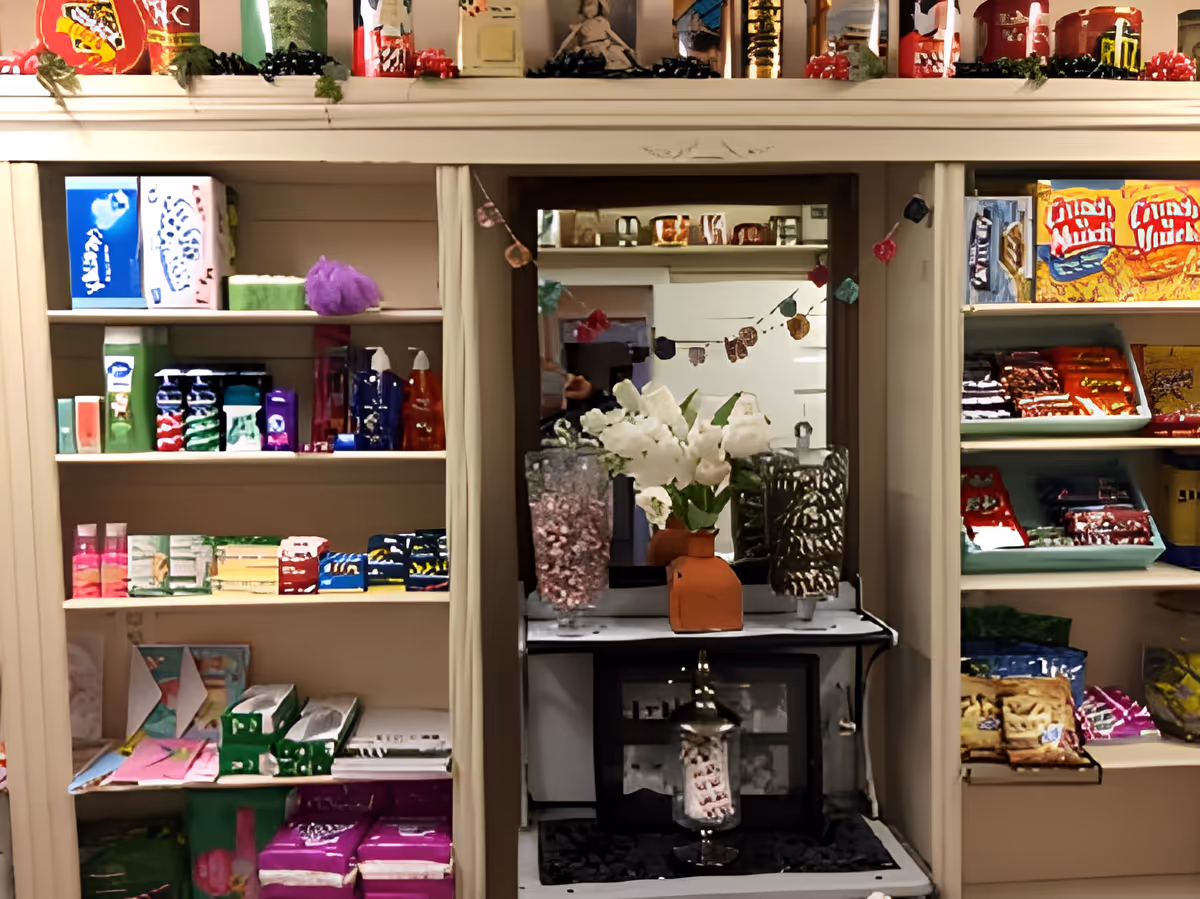 Interior view of a small shop or pantry area with shelves stocked with various packaged goods and toiletries. In the center, there is a mirror above a small counter holding a vase with white flowers and two large glass jars filled with candy or snacks. The shelves contain colorful boxes, bottles, and snack packages. A decorative garland hangs reflected in the mirror.