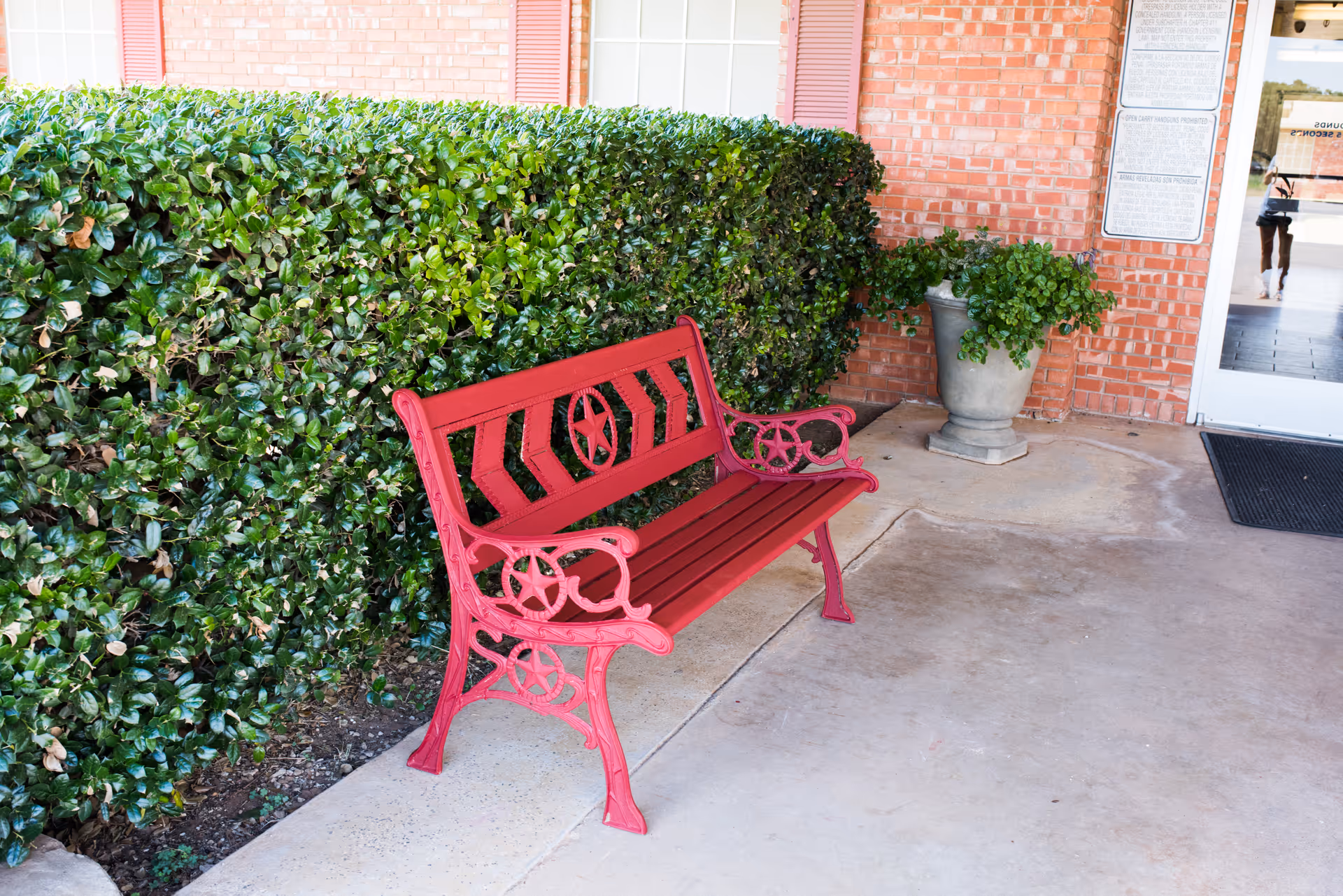 A red decorative bench on a concrete entrance patio next to trimmed hedges and a brick wall by the facility doorway.