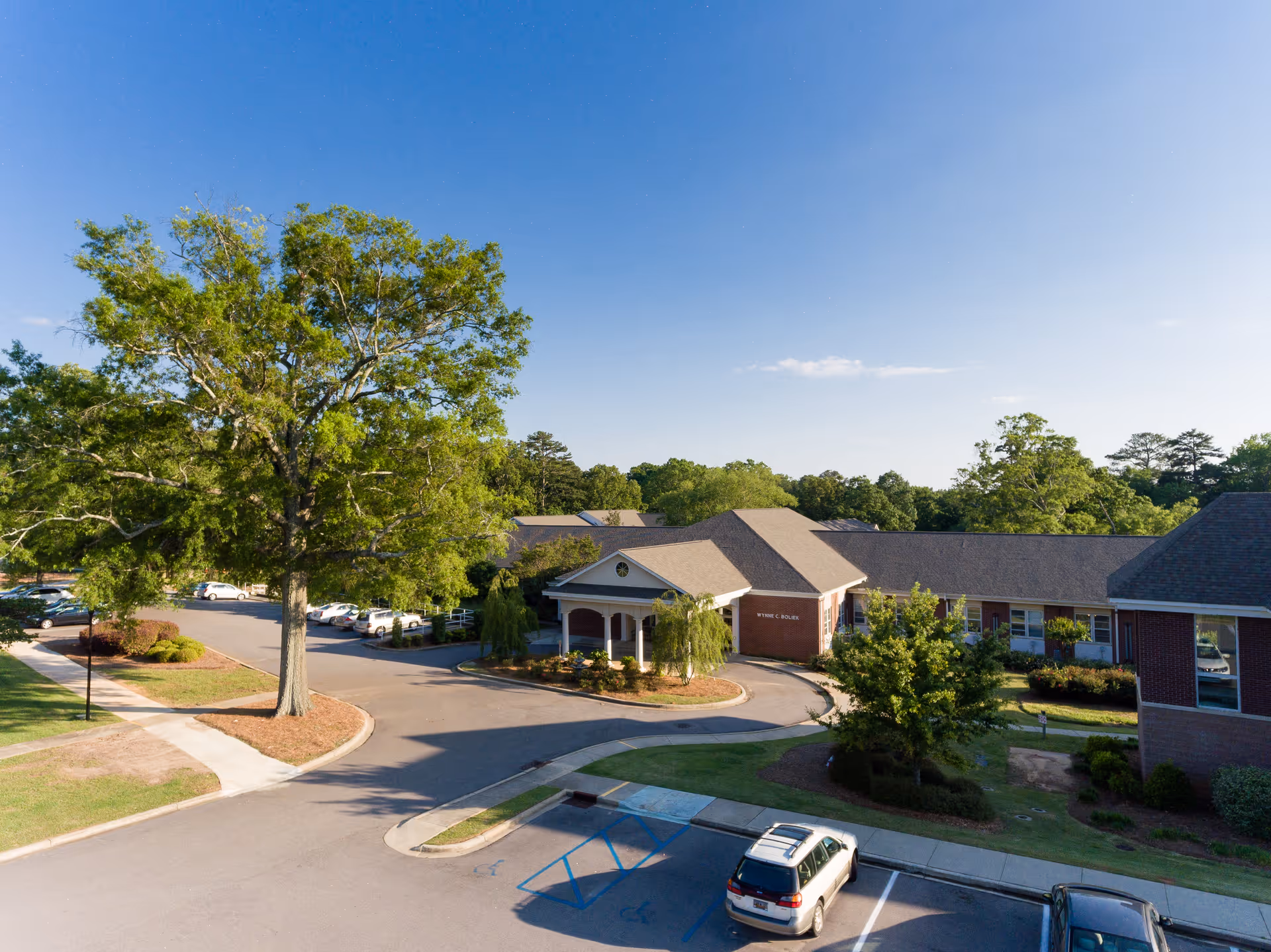 Exterior view of The Heritage At Lowman senior living facility showing a brick building with a covered entrance, surrounded by trees and a parking lot with several cars under a clear blue sky.