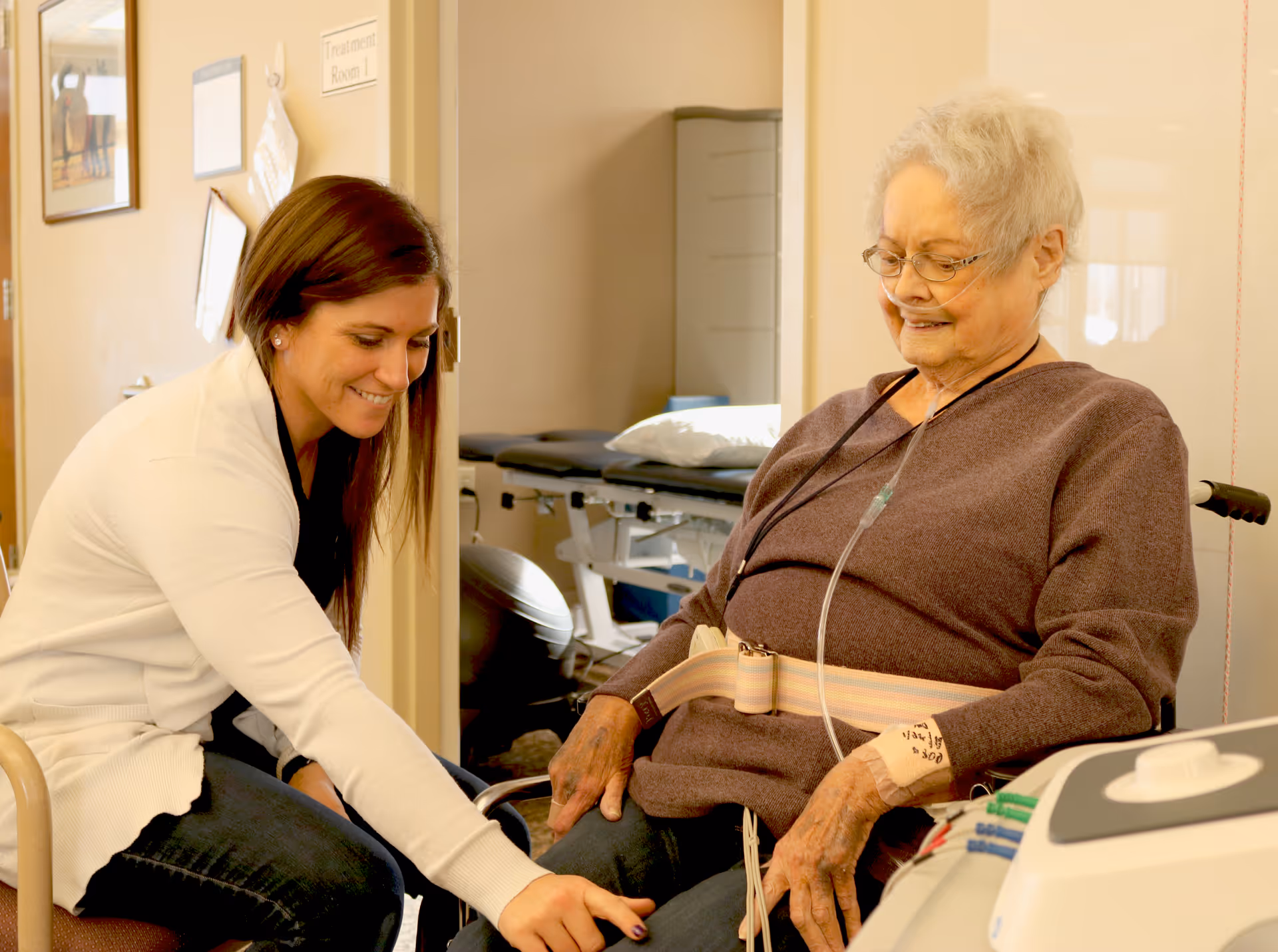 A healthcare professional is smiling and holding the hand of an elderly woman who is sitting in a wheelchair with an oxygen tube. They are in a medical treatment room with an examination table in the background.