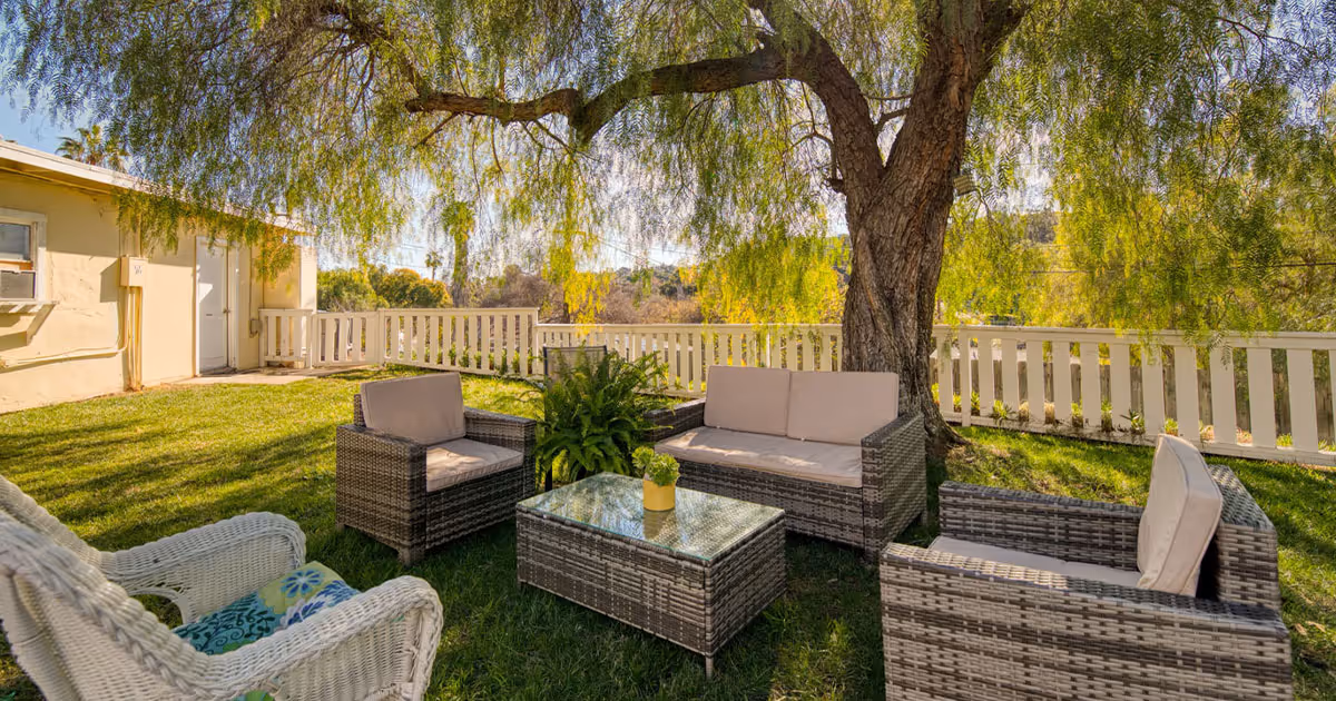 Outdoor seating area with wicker furniture including a sofa, two armchairs, and a glass-top coffee table arranged on a grassy lawn under a large tree with hanging branches. A white wicker chair with a patterned cushion is also visible. A white picket fence and a beige building with a door and window are in the background.