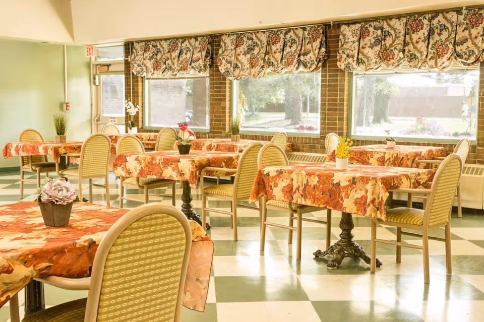 Bright dining room with tables covered in autumn-patterned tablecloths, chairs with woven backs, potted centerpieces, and large windows with floral valances.