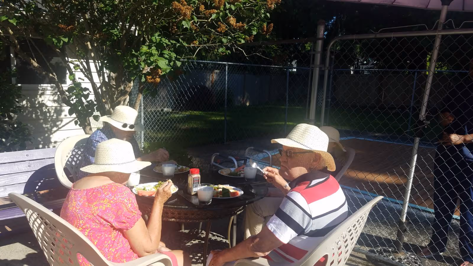 Four elderly people wearing sun hats sitting around a round outdoor table, eating a meal together in a fenced backyard with trees and grass visible in the background.