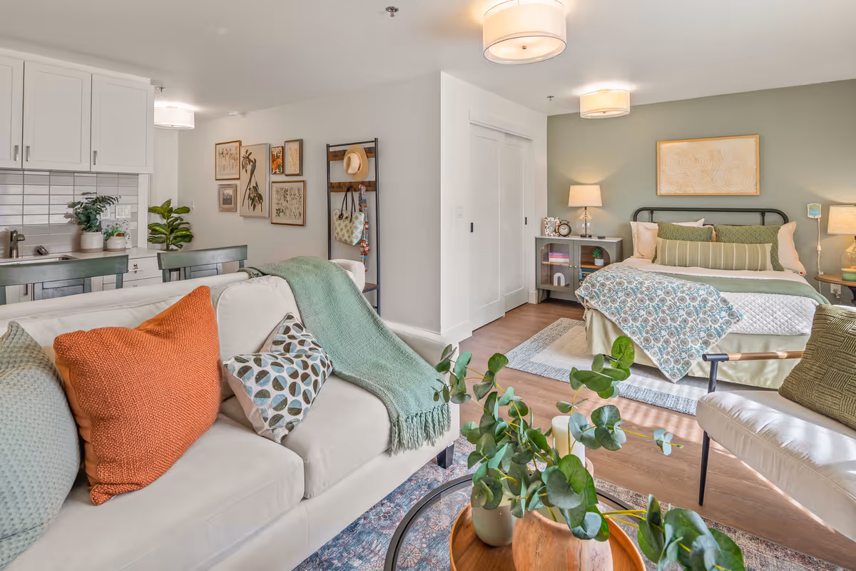A bright and cozy senior living apartment featuring a living area with a white sofa adorned with colorful pillows and a green throw blanket. In the background, there is a bedroom with a neatly made bed, bedside tables with lamps, and a green accent wall. The kitchen area with white cabinets and a tiled backsplash is partially visible to the left. The space is decorated with plants, framed artwork, and a coat rack with a hat and bag.