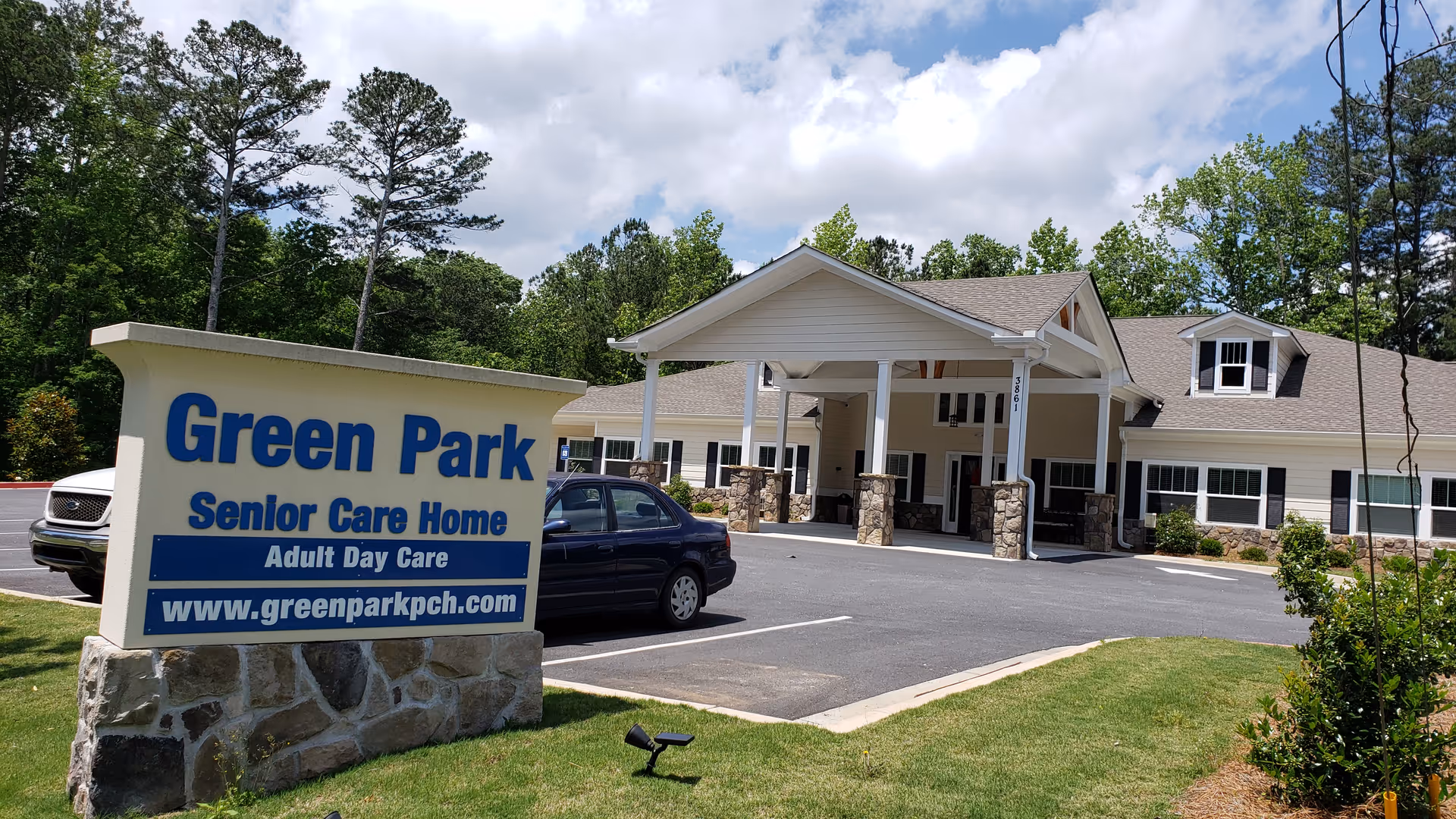 Exterior view of Green Park Senior Care Home building with a parking lot in front and a large sign displaying the facility name, services, and website. The building has a covered entrance supported by stone pillars and is surrounded by greenery and trees under a partly cloudy sky.