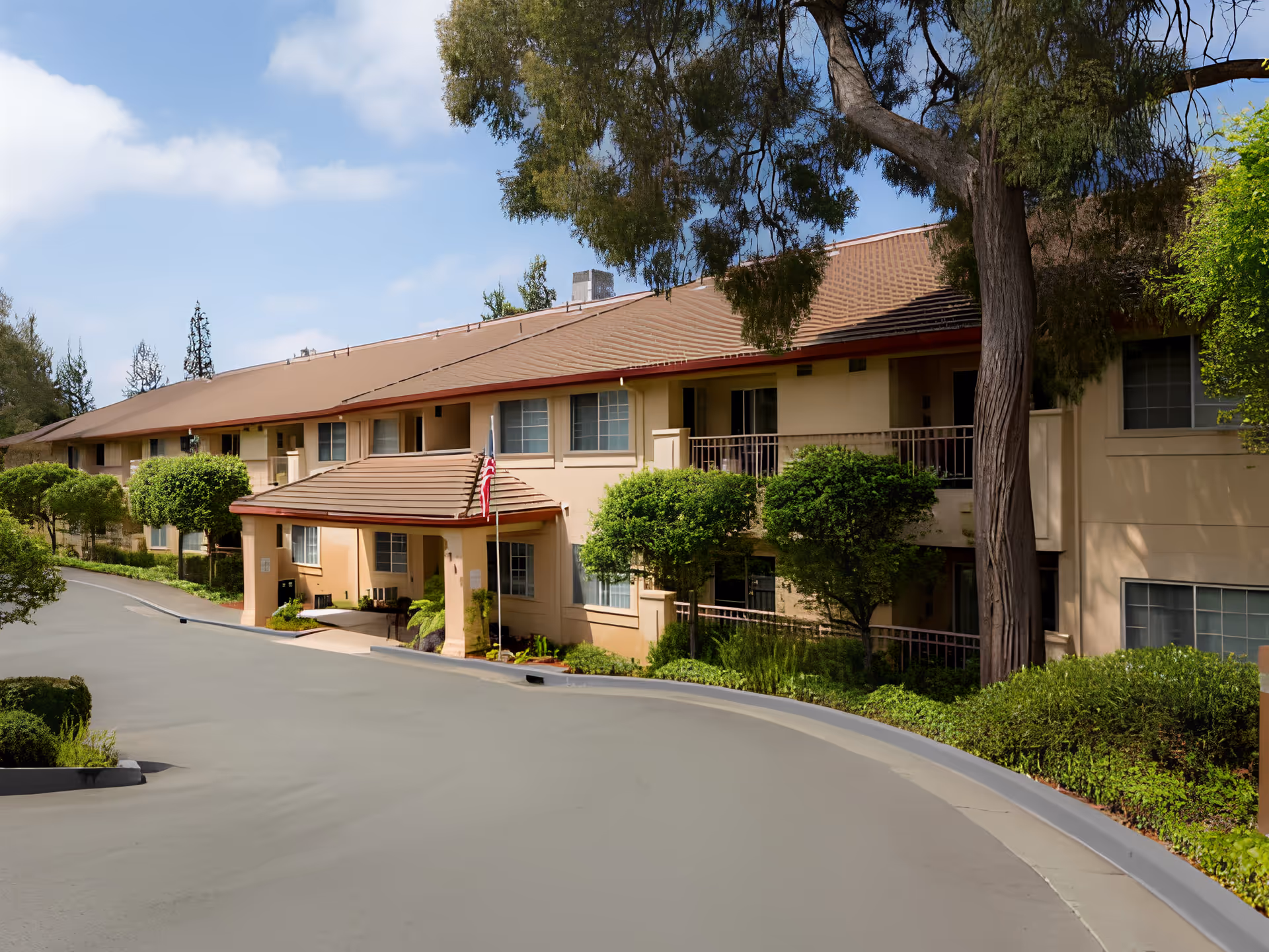 Exterior view of a two-story senior living facility building with beige walls and a brown roof. The entrance has a covered portico with an American flag. There are several windows and balconies, surrounded by green trees and bushes under a partly cloudy sky.