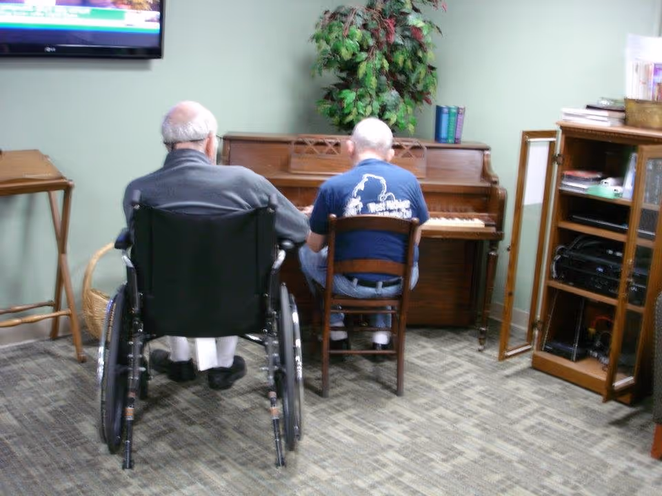 Two people seated in a communal room, one in a wheelchair and one at an upright piano.