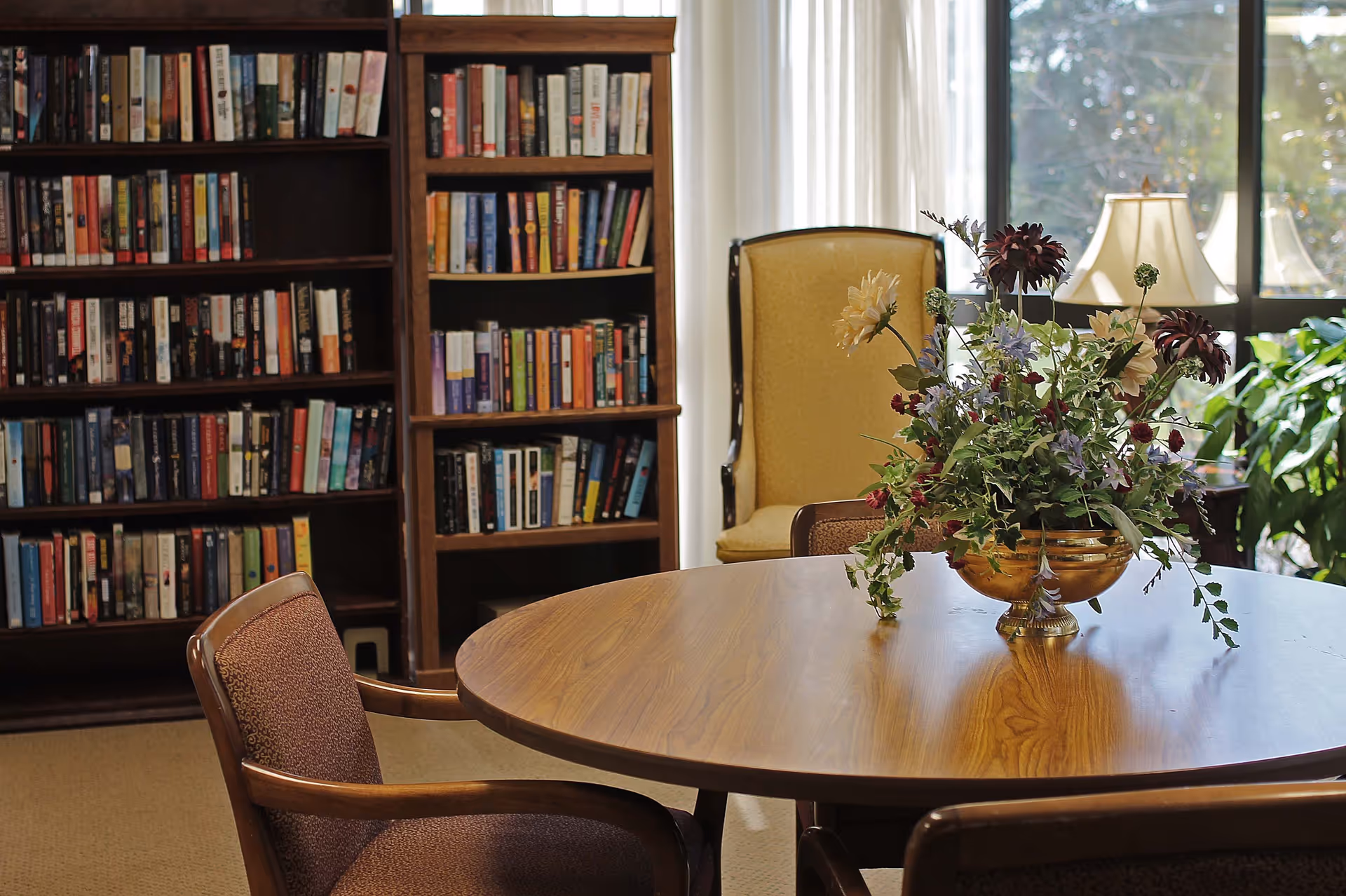 A cozy interior room with a round wooden table in the foreground, adorned with a decorative floral arrangement in a gold vase. Surrounding the table are upholstered chairs. In the background, there are bookshelves filled with books, a yellow armchair, a window with white curtains, a table lamp, and some green plants.