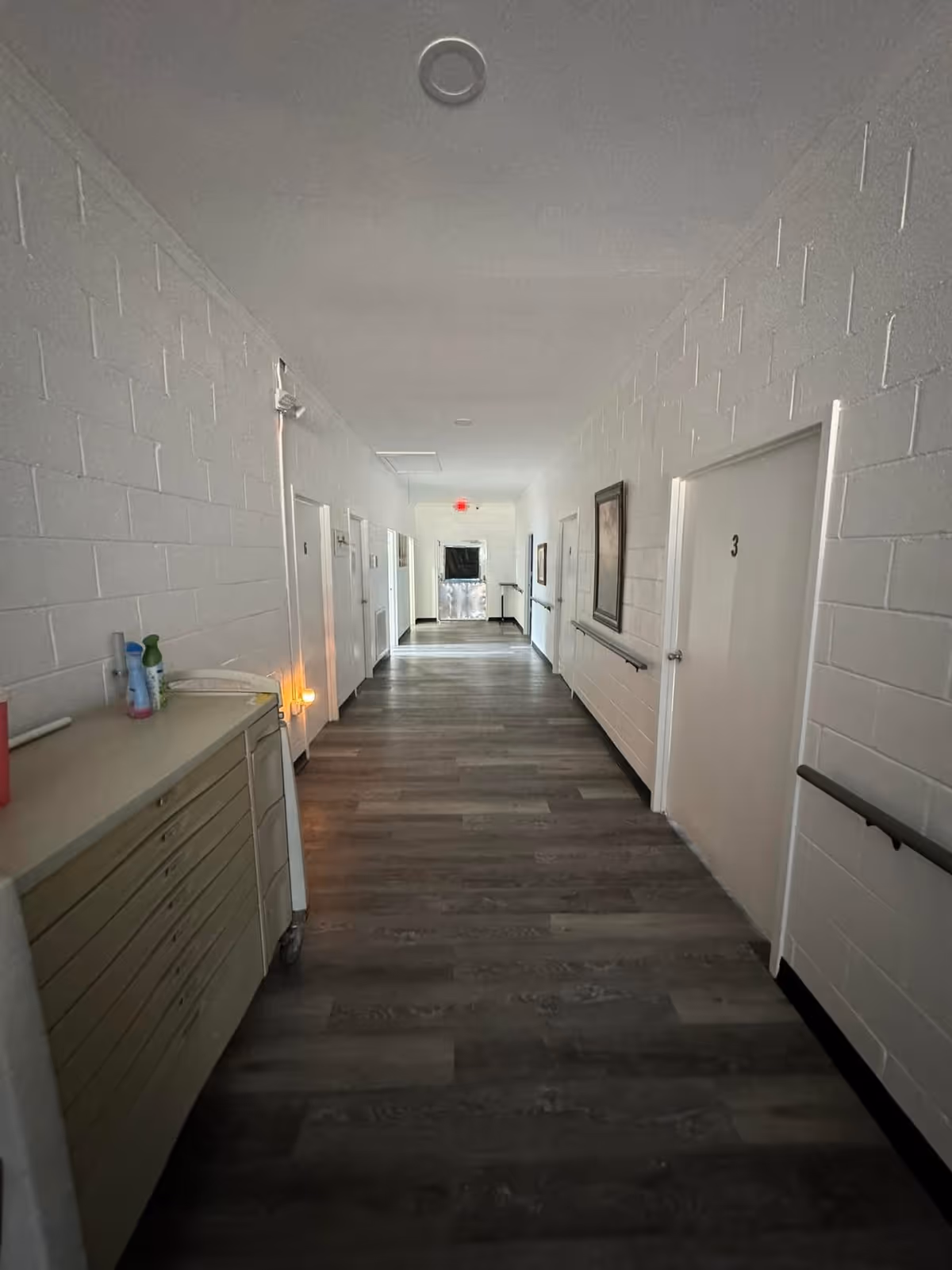 A long, narrow hallway in a facility with white brick walls, several closed white doors numbered 3 and 5, handrails on both sides, a cabinet with cleaning supplies on the left, and a framed picture on the right wall. The floor is covered with wood-patterned vinyl or laminate flooring, and there is a red exit sign visible at the far end.