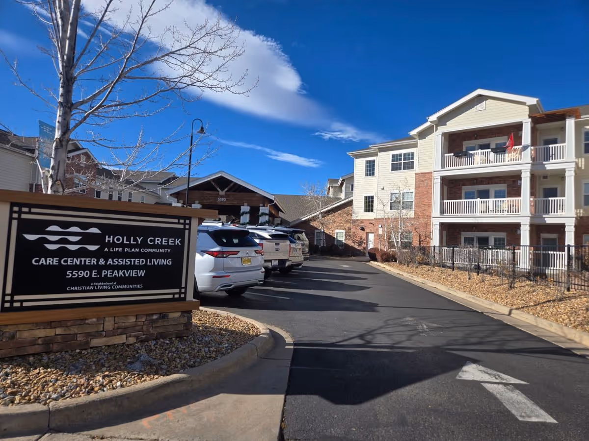 Exterior view of the Holly Creek assisted living facility showing the entrance drive, parked cars, and a sign in the foreground.