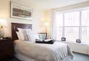 Bright, neatly made bedroom with a headboard, nightstands and lamps, artwork above the bed, and a large window with a view.