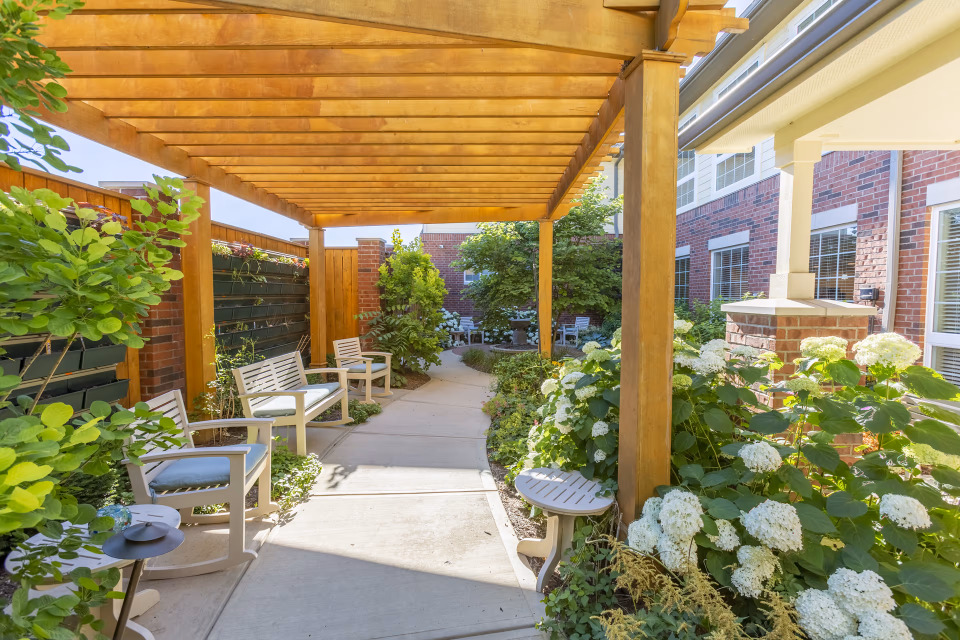 A sunny outdoor garden walkway at Independence Village of Zionsville West featuring a wooden pergola overhead, white cushioned chairs and benches along the path, lush green plants, and white flowering bushes. The path is paved and bordered by brick walls and building exterior.
