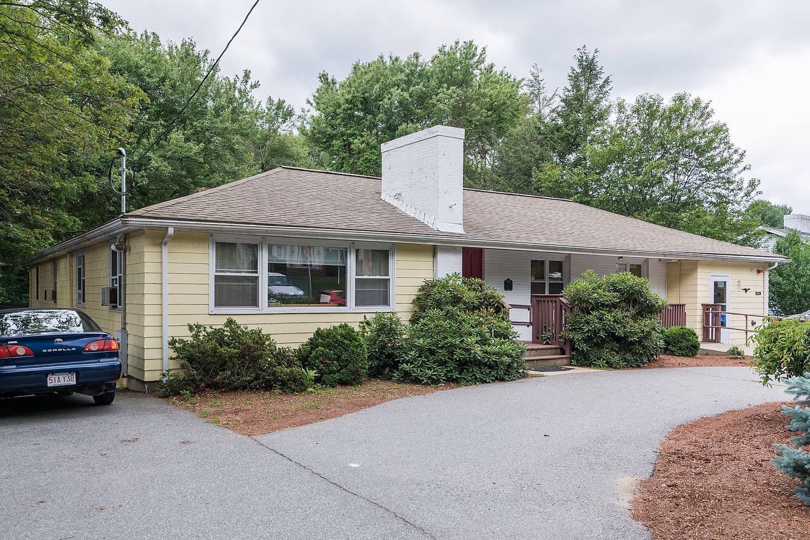 Single-story yellow house with a front porch, shrubs, driveway and a parked car set against a backdrop of trees.