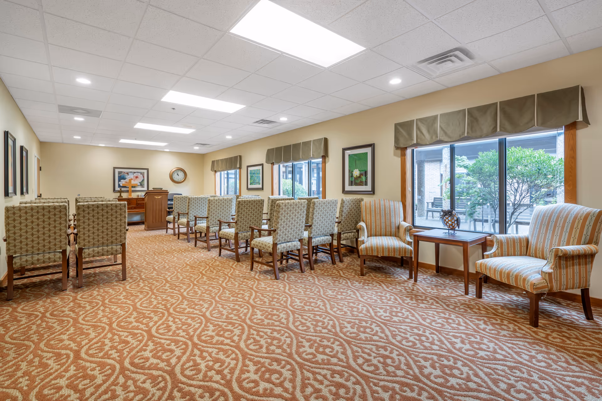 A well-lit room with patterned carpet and beige walls, featuring rows of upholstered chairs facing a wooden podium with a cross on it. The room has large windows with valances, framed artwork on the walls, and two striped armchairs next to a small wooden table with a decorative owl figurine. Outside the windows, greenery and outdoor seating are visible.