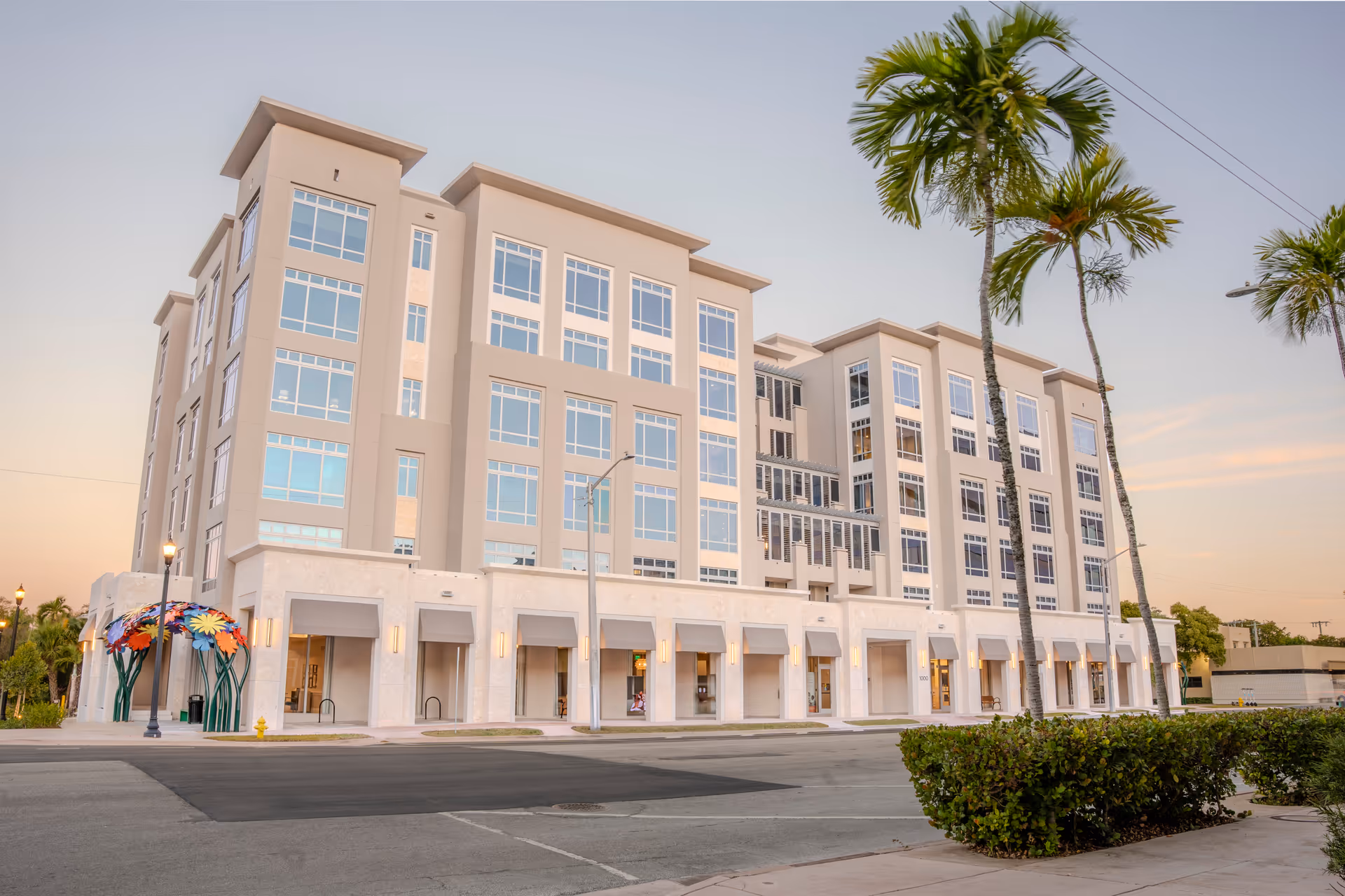 Large multi-story beige building with many windows, palm trees and a street in front under a pastel sky.