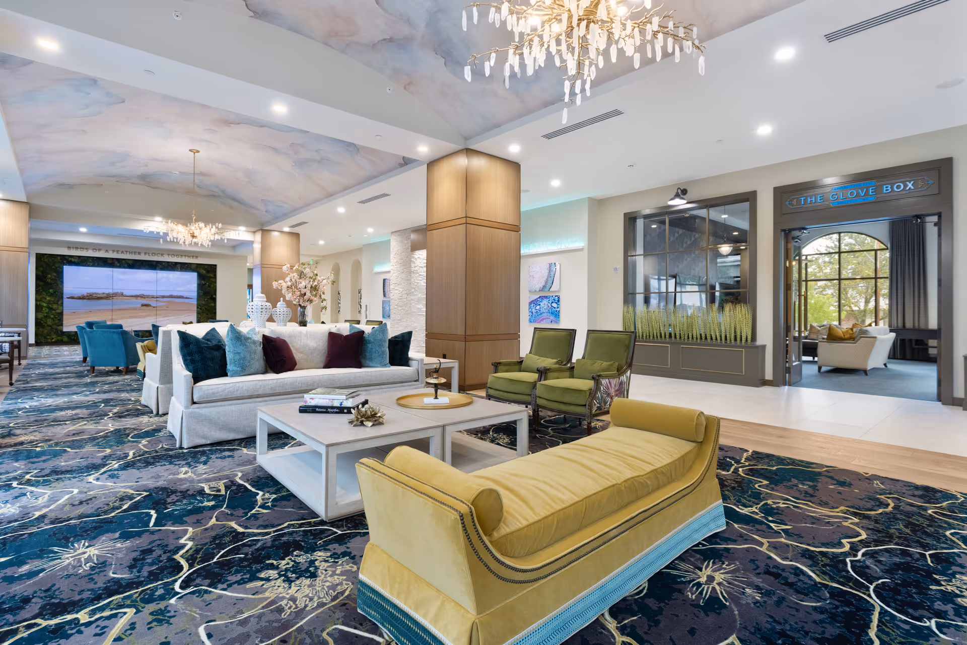 A spacious and elegantly decorated living room area in a senior living facility featuring a large patterned carpet, a yellow chaise lounge, green armchairs, a white sofa with colorful cushions, and a white coffee table with decorative items. The ceiling has a soft cloud-like mural and a modern chandelier. In the background, there is a large screen displaying a beach scene and a doorway labeled 'The Glove Box' leading to another seating area with large windows.