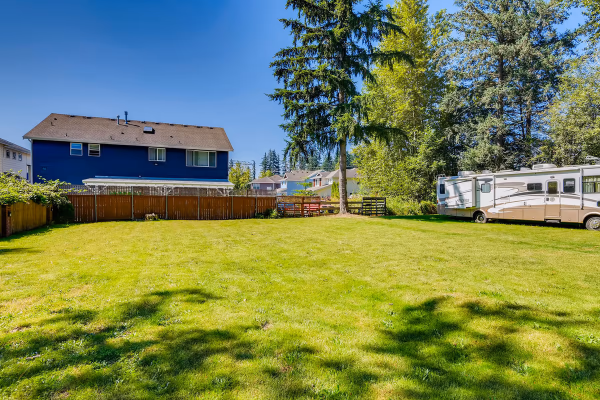 A spacious green lawn with a large tree in the middle, surrounded by a wooden fence. In the background, there is a two-story blue house and a large white and beige RV parked on the right side. Other houses and tall trees are visible beyond the fence under a clear blue sky.