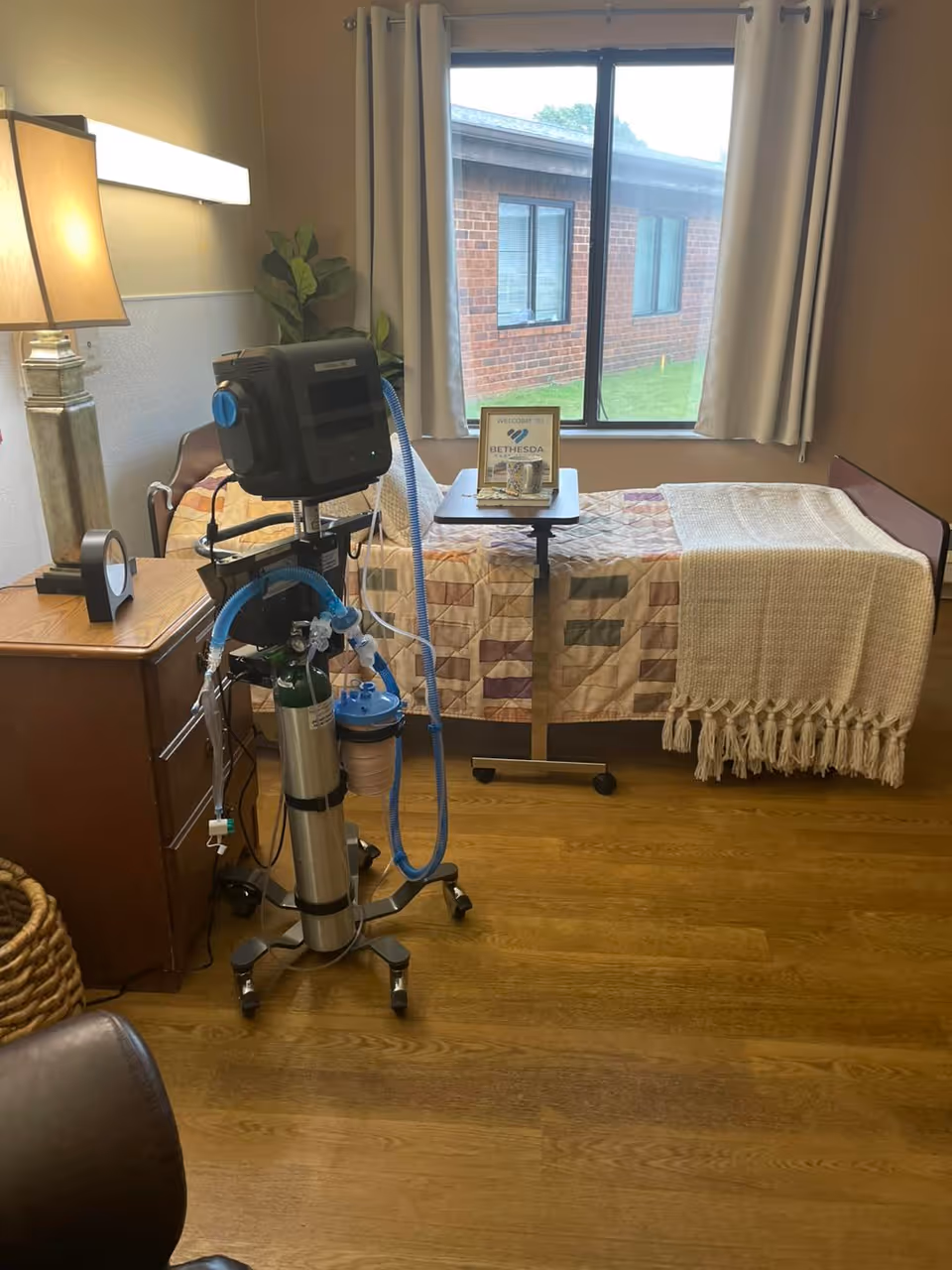 A small bedroom with a single bed covered with a patterned quilt and a beige throw blanket. Next to the bed is a wooden nightstand with a lamp and a clock. A medical oxygen machine with tubes is positioned near the bed. A window with beige curtains shows a view of a brick building outside. A small table on wheels is placed over the bed with a welcome sign for Bethesda Care Center.