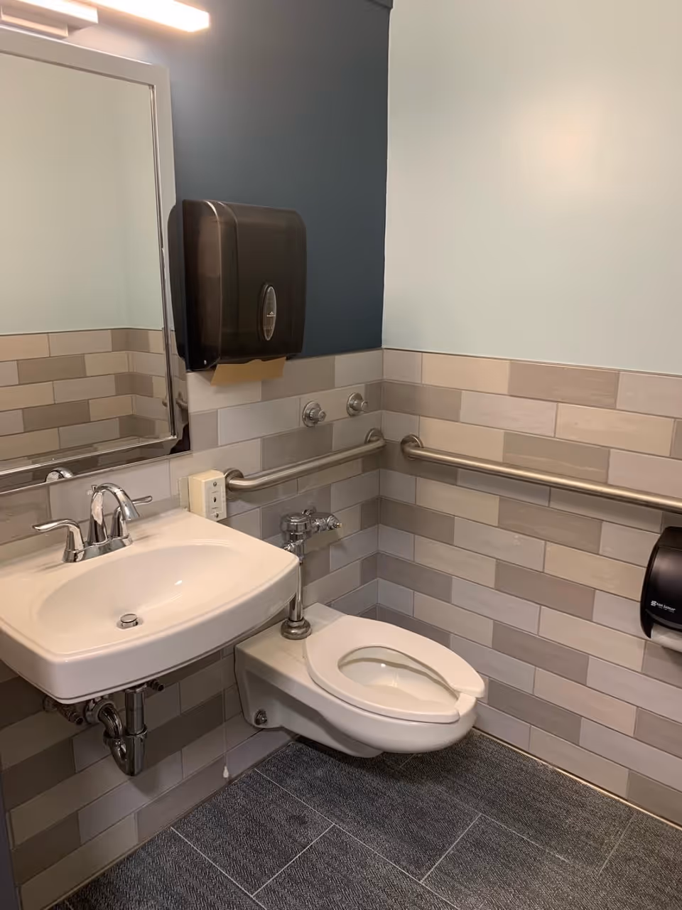 A clean and modern bathroom with a wall-mounted toilet and a white sink. The walls are tiled with beige and light gray rectangular tiles, and there are stainless steel grab bars installed around the toilet for accessibility. A large mirror is above the sink, and a black paper towel dispenser is mounted on the wall.