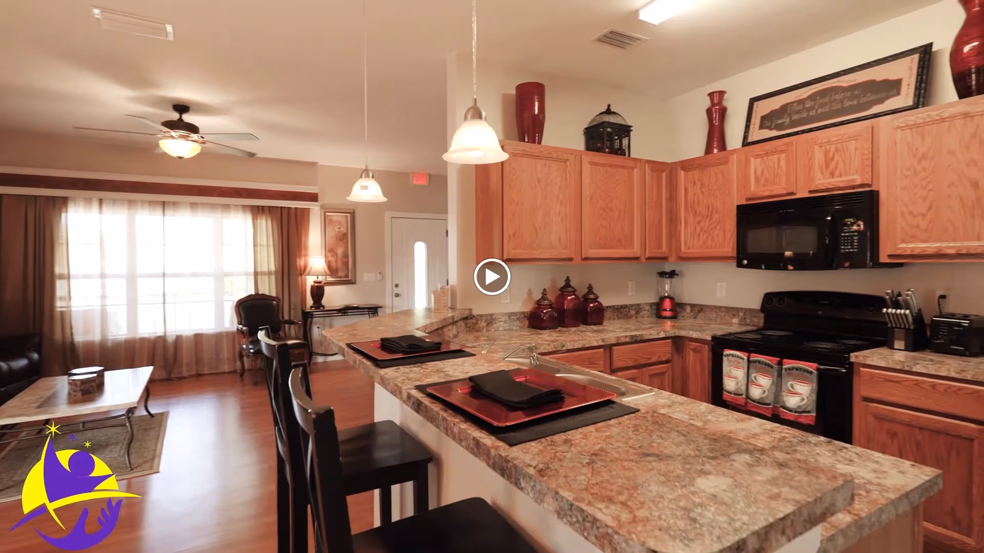 Open-plan kitchen with a marble countertop island and bar stools opening to a living area with curtains and a ceiling fan.