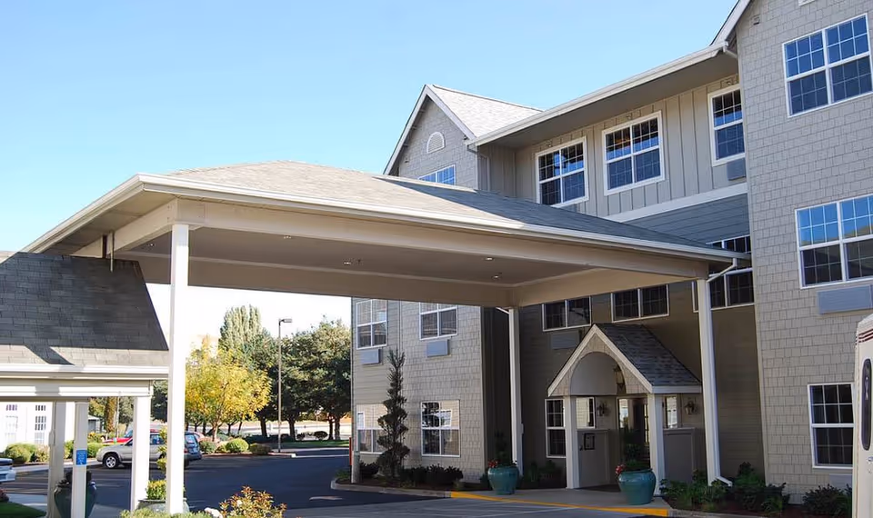 Main entrance of a senior living building with a covered drop-off canopy, potted plants, and a paved driveway.
