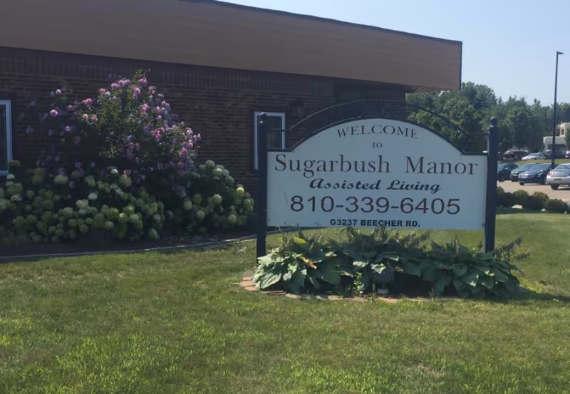 Outdoor view of the entrance area of Sugarbush Manor assisted living facility, featuring a sign with the facility's name, phone number, and address, surrounded by green grass and flowering bushes in front of a brick building.