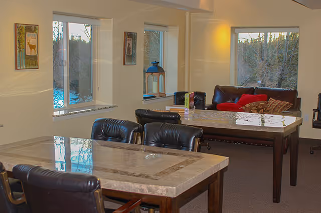 Interior view of a common area in Aspen Creek - Riverside Assisted Living featuring two marble-topped tables with black leather chairs around them. In the background, there is a dark brown leather couch with red and patterned pillows near windows showing an outdoor wooded area. The walls are light-colored with framed artwork and a decorative lantern on the windowsill.