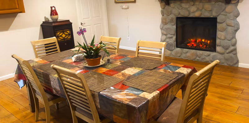 A dining room with a rectangular table covered by a colorful patchwork tablecloth and six wooden chairs around it. A potted orchid plant is placed in the center of the table. In the background, there is a stone fireplace with a glowing fire and a small wooden cabinet with a decorative item on top. The floor is wooden, and the walls are light-colored.