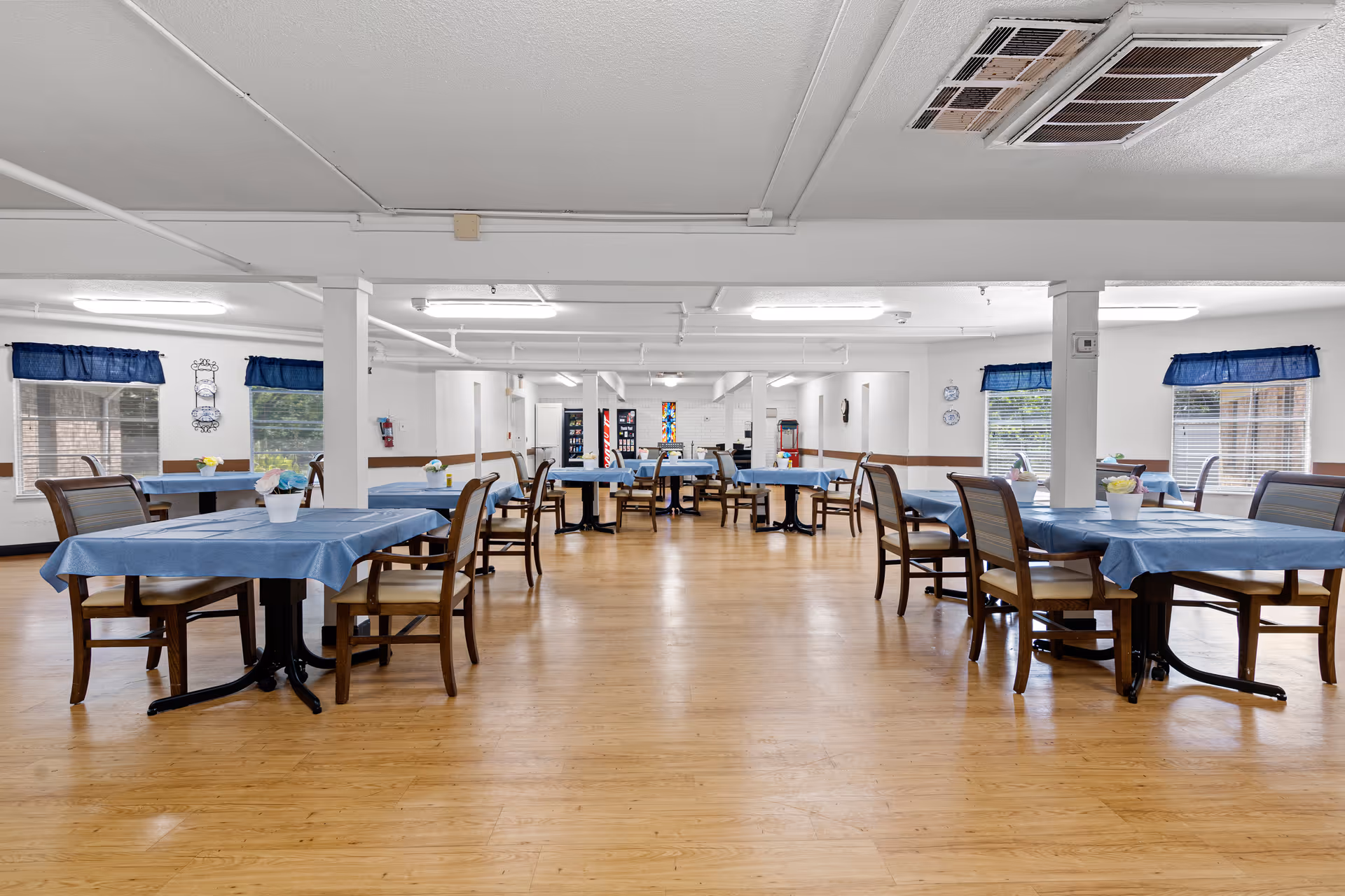 A spacious dining room with multiple tables covered in blue tablecloths, each surrounded by wooden chairs with cushioned seats. The room has large windows with blue valances, light wood flooring, white walls, and ceiling lights. Small flower pots are placed on each table.