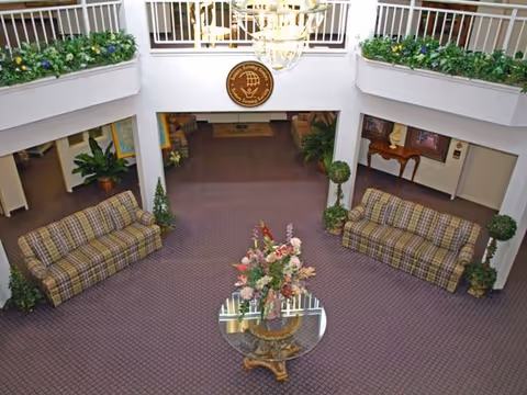 Interior view of a senior living facility lobby with two plaid sofas facing each other, a round glass table with a floral arrangement in the center, potted plants, and a chandelier hanging from the ceiling. There is a balcony with white railings and greenery above.