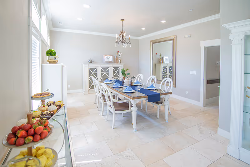 Bright, elegant dining room with a long set table, white chairs, a chandelier, large mirror, and a side table with fruit and pastries.