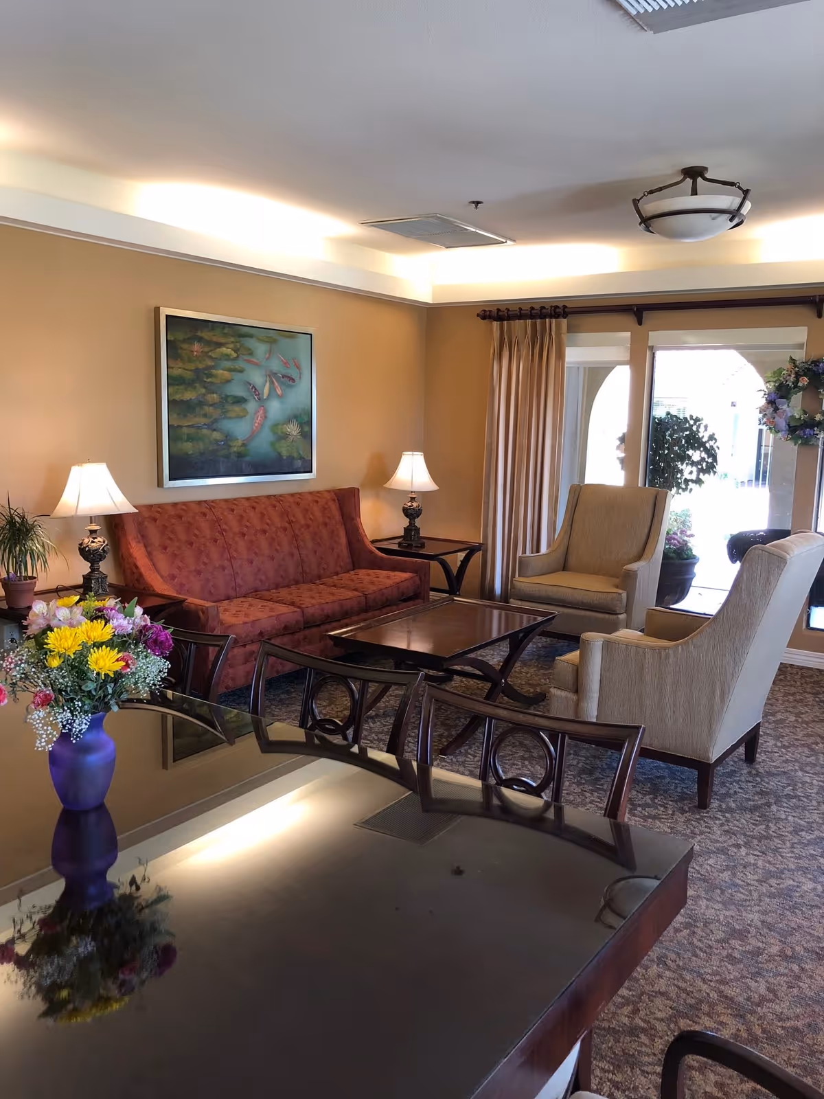 A cozy senior living common area with a red patterned sofa, two beige armchairs, a wooden coffee table, and side tables with lamps. There is a large window with beige curtains and a glass door decorated with a floral wreath. A framed painting of koi fish and lily pads hangs on the tan wall. A dark wooden table with chairs and a purple vase with colorful flowers is in the foreground.
