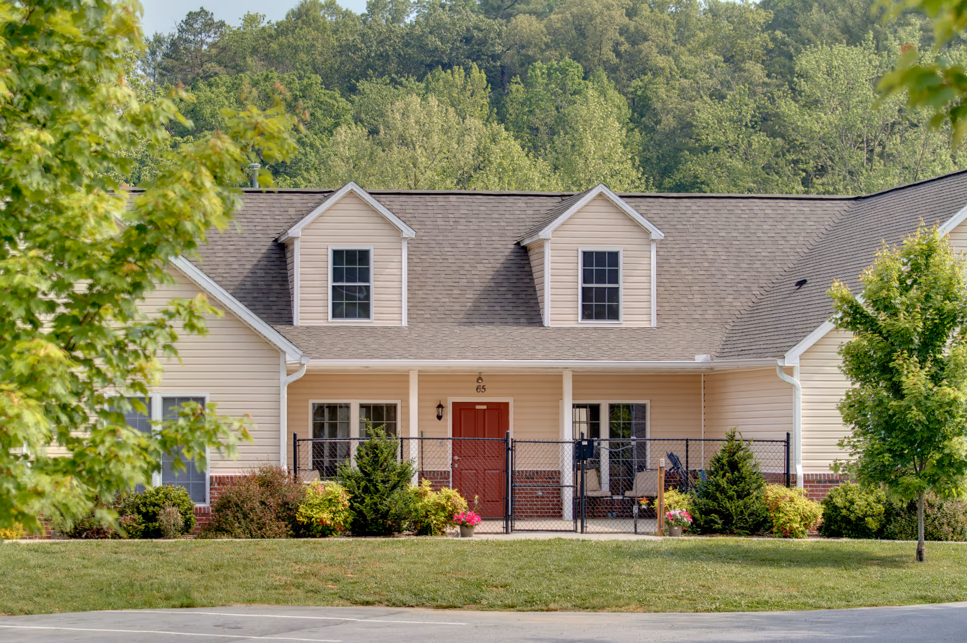 Front exterior view of a single-story residential building with beige siding, a brown shingled roof, two dormer windows, a red front door, and a fenced porch area. The building is surrounded by green grass, bushes, and trees with a forested area in the background.